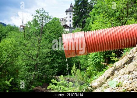 Un jet d'eau mince s'écoule d'un tuyau d'égout en plastique rouge contre un fond flou d'arbres et d'un bâtiment Banque D'Images