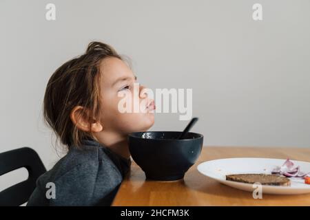 Une fillette malheureuse mange de la soupe dans un bol noir avec du pain et de l'oignon.Photo de style de vie d'un enfant dans la cuisine ayant un repas, hurlant enfant.Mangeur difficile Banque D'Images