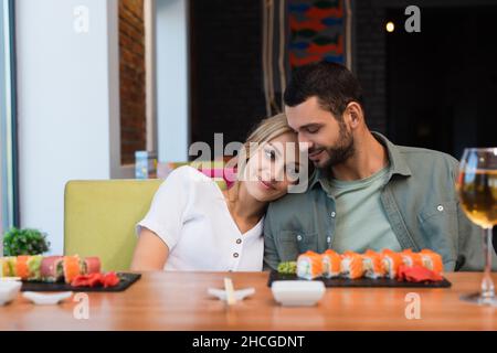 jeune et heureux couple assis près de petits pains à sushis flous et d'un verre de vin blanc au restaurant Banque D'Images