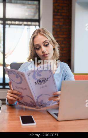jeune femme blonde lisant le menu près d'un ordinateur portable et d'un smartphone avec écran vierge Banque D'Images