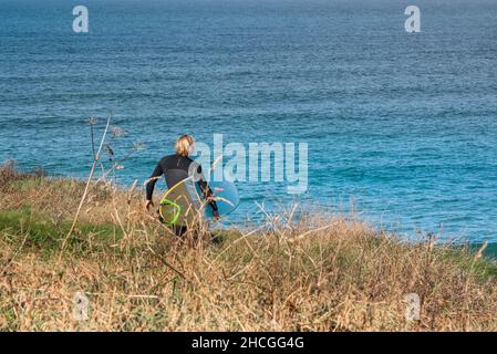 Un surfeur mâle transportant sa planche de surf marchant sur un sentier côtier jusqu'à la mer dans les Cornouailles. Banque D'Images