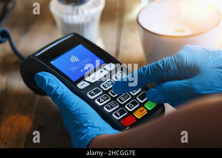 La main d'une femme avec des gants de protection en latex médical tient le terminal de paiement et saisit le code PIN dans un café.Protection contre le coronavirus ou COVID-19. Banque D'Images