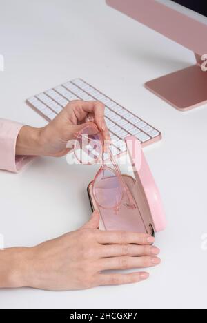 Angleterre, Royaume-Uni.2021. Les mains des femmes enlever les lunettes de l'étui avec un fond d'ordinateur et de clavier, tous roses. Banque D'Images