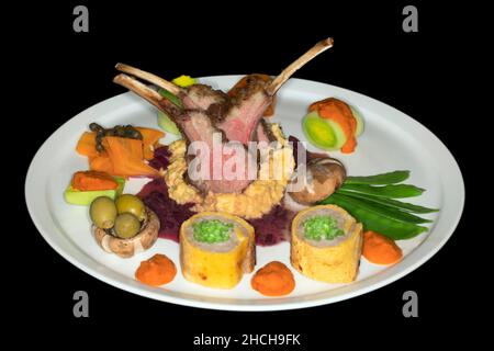 Selle d'agneau rôtie rose sur polenta de maïs avec oignons rouges, sauce au persil et légumes variés, photographie en studio avec fond noir Banque D'Images