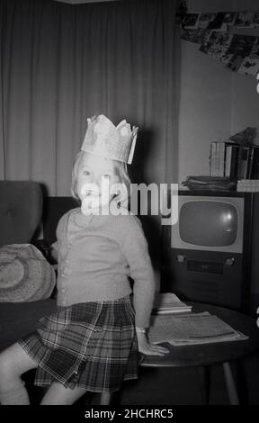 Fin 1950s, une jeune fille portant un chapeau de papier de papillote de noël, posant pour une photo dans le salon, devant un téléviseur de l'époque, Angleterre, Royaume-Uni. Banque D'Images