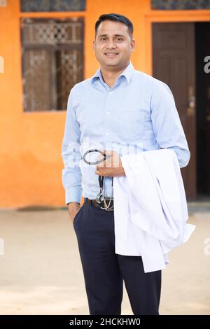 Portrait de jeune homme indien souriant Docteur portant une chemise tenant le stéthoscope et le tablier, il regarde la caméra avec des émotions positives contre l'orang Banque D'Images