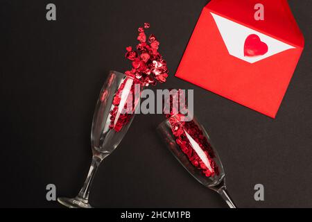 vue de dessus des verres à champagne avec coeurs confetti rouges près de l'enveloppe sur noir Banque D'Images