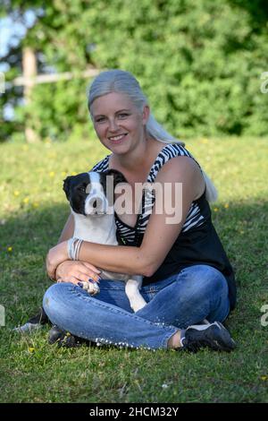 Une jeune femme blonde est assise sur une pelouse fraîchement verte et embrasse et câlin son chien. Banque D'Images