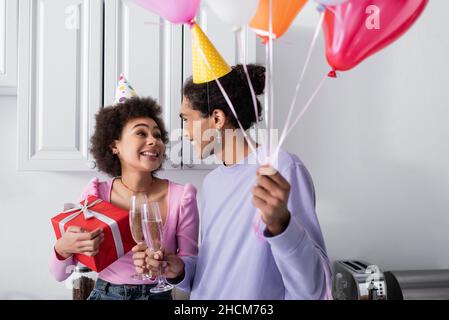 Femme afro-américaine positive tenant le présent et toaster le champagne avec un petit ami tenant des ballons à la maison Banque D'Images