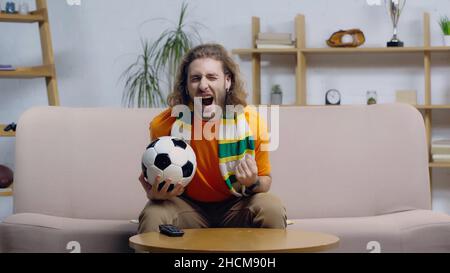 homme enthousiaste tenant le ballon de football et montrant le geste de triomphe tout en regardant le championnat sur la télé à domicile Banque D'Images