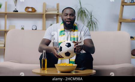 un homme afro-américain enthousiaste tenant le ballon de football tout en regardant le championnat de football près de bière et de jetons Banque D'Images