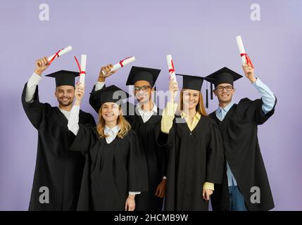 Heureux diplômés d'université multiraciale en casquettes et robes de chambre tenant des diplômes et souriant Banque D'Images