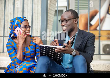 belle femme d'affaires mûre en robe traditionnelle assise à l'extérieur sur les escaliers parlant sur le téléphone portable tandis que sa collègue rit à elle. Banque D'Images