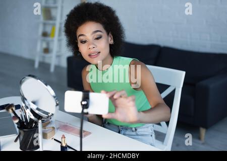 Blogueur afro-américain utilisant un smartphone flou près de cosmétiques décoratifs à la maison Banque D'Images