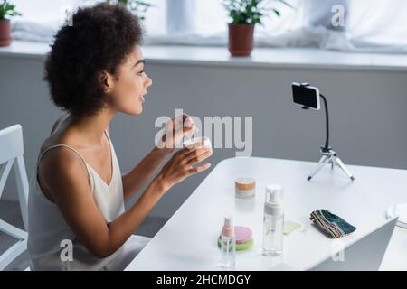 Vue latérale d'un blogueur afro-américain souriant tenant une jarre avec des cellules oculaires près d'un smartphone à la maison Banque D'Images