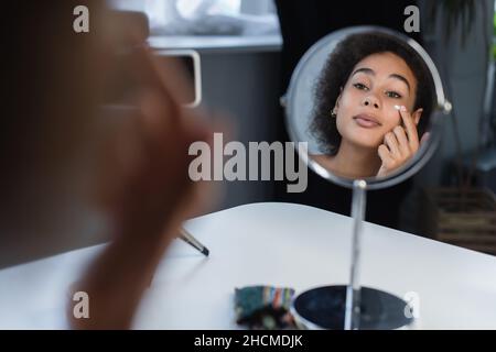 Blogueur afro-américain appliquant de la crème cosmétique sur le visage près du miroir et du smartphone à la maison Banque D'Images