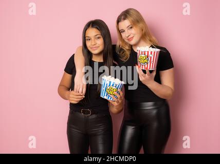 Deux filles amies en noir posant ensemble avec du maïs pop sur fond rose. Banque D'Images