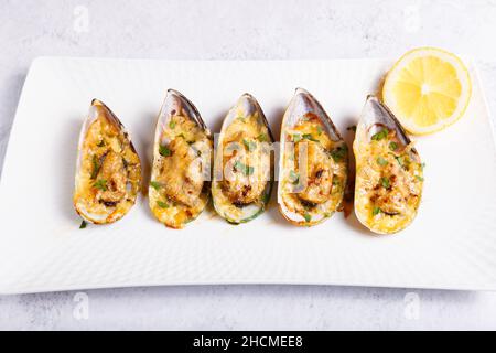 Moules de coquille de bœuf cuites dans une demi-coquille avec fromage, persil et citron sur une assiette blanche. Hors-d'œuvre chaud traditionnel. Gros plan, vue de dessus. Banque D'Images