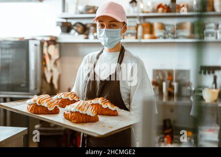 L'employé dans le masque tient le plateau avec des croissants frais décorés dans la boulangerie Banque D'Images