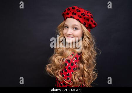 Suivant son style personnel.Petite fille dans un chapeau de style français.Bonne fille avec de longs cheveux bouclés dans un béret rouge et une robe.Enfant parisien sur fond noir.La mode et la beauté de l'été. Banque D'Images