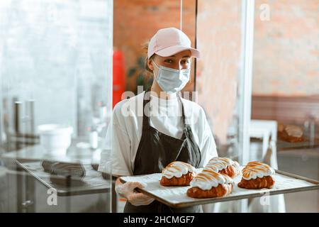 Femme dans le masque tient le plateau avec des croissants décorés debout dans la boulangerie artisanale Banque D'Images