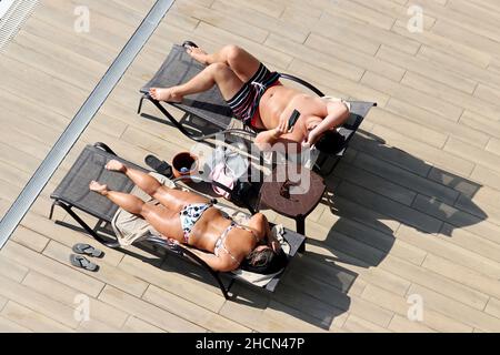 Couple allongé dans des chaises longues et bains de soleil près de la piscine, vue de dessus.Une femme se détend en maillot de bain et un homme en malles en utilisant un smartphone sur une plage Banque D'Images