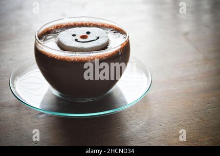 Un chocolat chaud dans une tasse en verre avec soucoupe et un guimauve à visage bonhomme de neige Banque D'Images