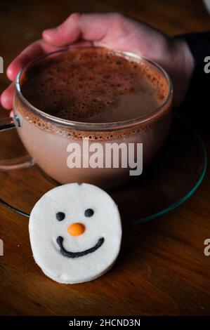 La main d'un enfant berce un chocolat chaud dans une tasse en verre avec soucoupe et un guimauve à visage bonhomme de neige Banque D'Images