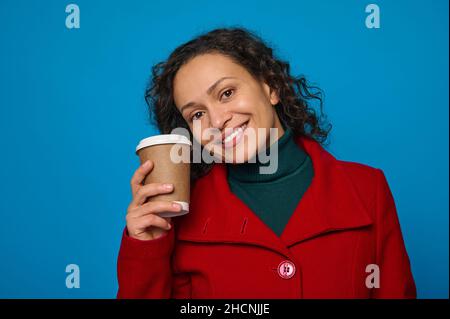 Portrait d'une charmante jolie femme aux cheveux sombres en pull vert et manteau rouge vif tient une tasse jetable en carton avec boisson chaude et sourires gaifu Banque D'Images