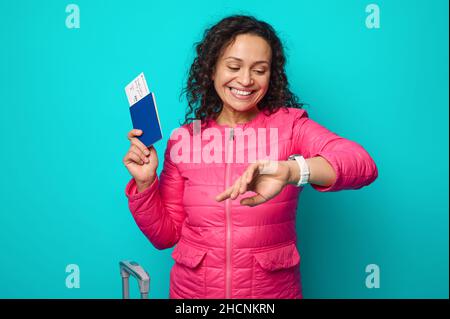 Jolie femme aux cheveux sombres, jolie femme tenant un passeport et une carte d'embarquement souriant regardant la montre, vérifiant l'heure, isolée sur fond bleu avec humour Banque D'Images