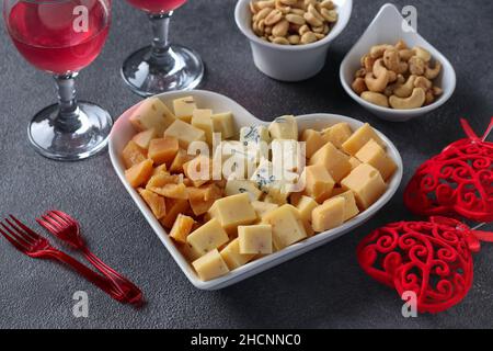 Variétés de fromage d'élite dans une assiette en forme de coeur, noix de cajou, arachides et deux verres de vin pour la Saint-Valentin sur fond gris Banque D'Images