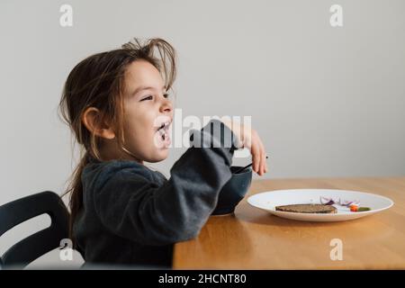 Une fillette malheureuse mange de la soupe dans un bol noir avec du pain et de l'oignon.Photo de style de vie d'un enfant dans la cuisine ayant un repas, hurlant enfant.Mangeur difficile Banque D'Images