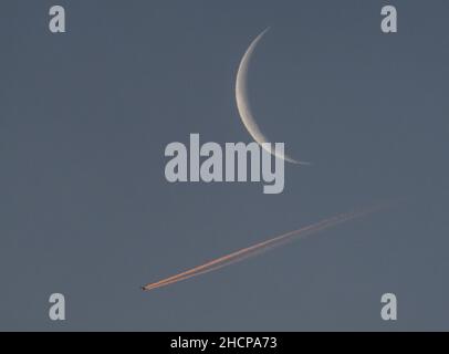 31 décembre 2021, Hessen, Francfort-sur-le-main: Un avion passe devant le croissant étroit de la lune en déclin au-dessus de Francfort-sur-le-main dans la matinée, son contrail illuminé par la lumière rougeâtre du soleil levant.Neuf pour cent du satellite terrestre est encore visible en ce moment, qui est actuellement à environ 363 000 kilomètres de la Terre.Photo: Frank Rumpenhorst/dpa Banque D'Images