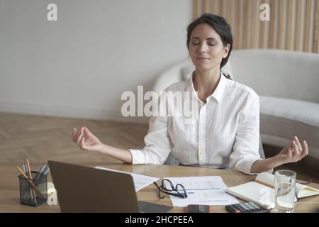 Femme italienne calme aux yeux fermés méditant sur le lieu de travail tout en étant assise à la table avec un ordinateur portable Banque D'Images