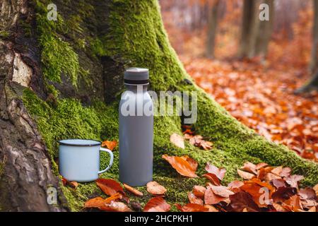 Thermos et mug de voyage en forêt.Contenant à boisson isolé.Tronc d'arbre moussy et feuilles d'automne tombées.Rafraîchissements pendant la randonnée dans la nature Banque D'Images