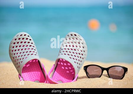 Gros plan sur les chaussures Clogs et les lunettes de soleil noires protectrices sur la plage de sable au bord de la mer tropicale par temps chaud et ensoleillé. Concept de vacances d'été. Banque D'Images