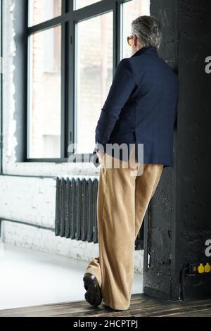 Vue arrière de l'homme d'âge moyen élégant en affaires vêtements décontractés et lunettes de soleil se penchant sur le mur, en regardant loin en posant dans l'intérieur de loft Banque D'Images