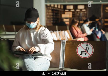 Une jeune femme dans un masque tire un ordinateur portable d'une couverture tout en étant assise sur des chaises avec des panneaux « Keep distance » au terminal de l'aéroport Banque D'Images