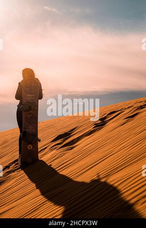 Femme avec planche à sable debout sur des dunes de sable dans le désert contre ciel nuageux Banque D'Images