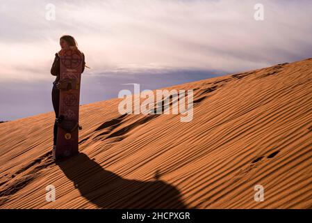 Femme avec planche à sable debout sur des dunes de sable dans le désert contre ciel nuageux Banque D'Images