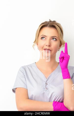 Portrait d'une jeune femme médecin de race blanche portant des gants en caoutchouc en uniforme médical avec un doigt vers le haut, regardant sur un fond blanc, espace de copie Banque D'Images