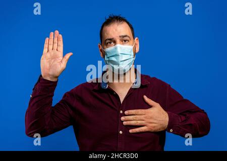 Portrait d'un grave homme brunette avec un masque médical chirurgical debout avec un bras relevé, jurant allégeance, prêtant serment d'expression responsable Banque D'Images