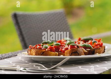 Hors-d'œuvre savoureux de tomates italiennes, ou bruschetta, sur des tranches de baguette grillée garnie de basilic, gros plan sur une assiette sur une table à l'extérieur Banque D'Images