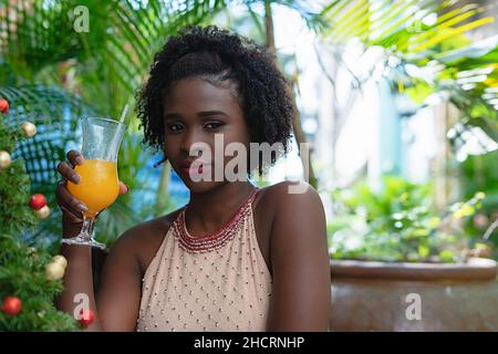 jeune femme latine noire en robe élégante assise à l'extérieur en buvant une boisson rafraîchissante Banque D'Images