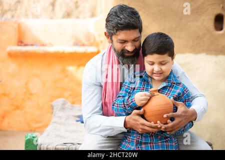 Père et fils indiens ruraux mettant la pièce dans la boîte d'argent en argile.Joyeux homme et garçon de barbe tenant une banque de porc traditionnelle ou Gullak, papa enseignant enfant à sa Banque D'Images