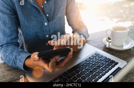 Jeune femme d'affaires vêtue de jeans assis à table dans le café et écrivant au smartphone.Sur la table se trouve un ordinateur portable et une tasse de café.Indépendant travaillant en collaboration Banque D'Images