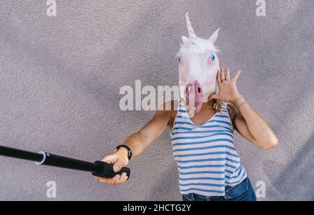 Une jeune femme figue dans un masque unicorn se tient sur un mur gris et utilise un bâton de selfie pour prendre des photos. Banque D'Images