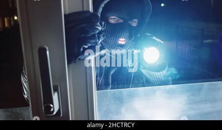 Burglar masqué regardant à travers la fenêtre avec une lampe de poche la nuit Banque D'Images