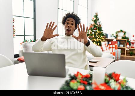 Jeune africain américain utilisant un ordinateur portable assis sur la table par arbre de noël peur et terrifié avec l'expression de peur stop geste avec les mains, shou Banque D'Images
