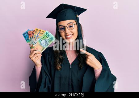 Jeune femme hispanique portant l'uniforme de la remise des diplômes tenant des dollars australiens souriant souriant avec la main et le doigt Banque D'Images